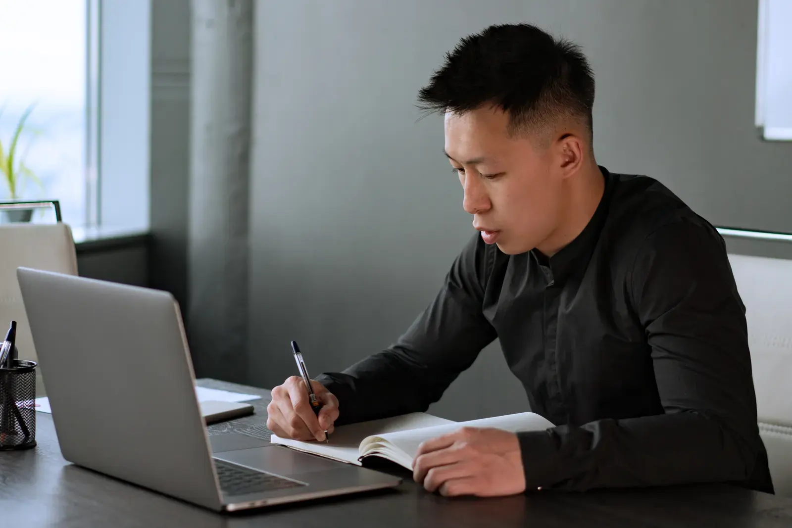 A professional man writes notes in a notebook at his desk with a laptop, demonstrating work focus.