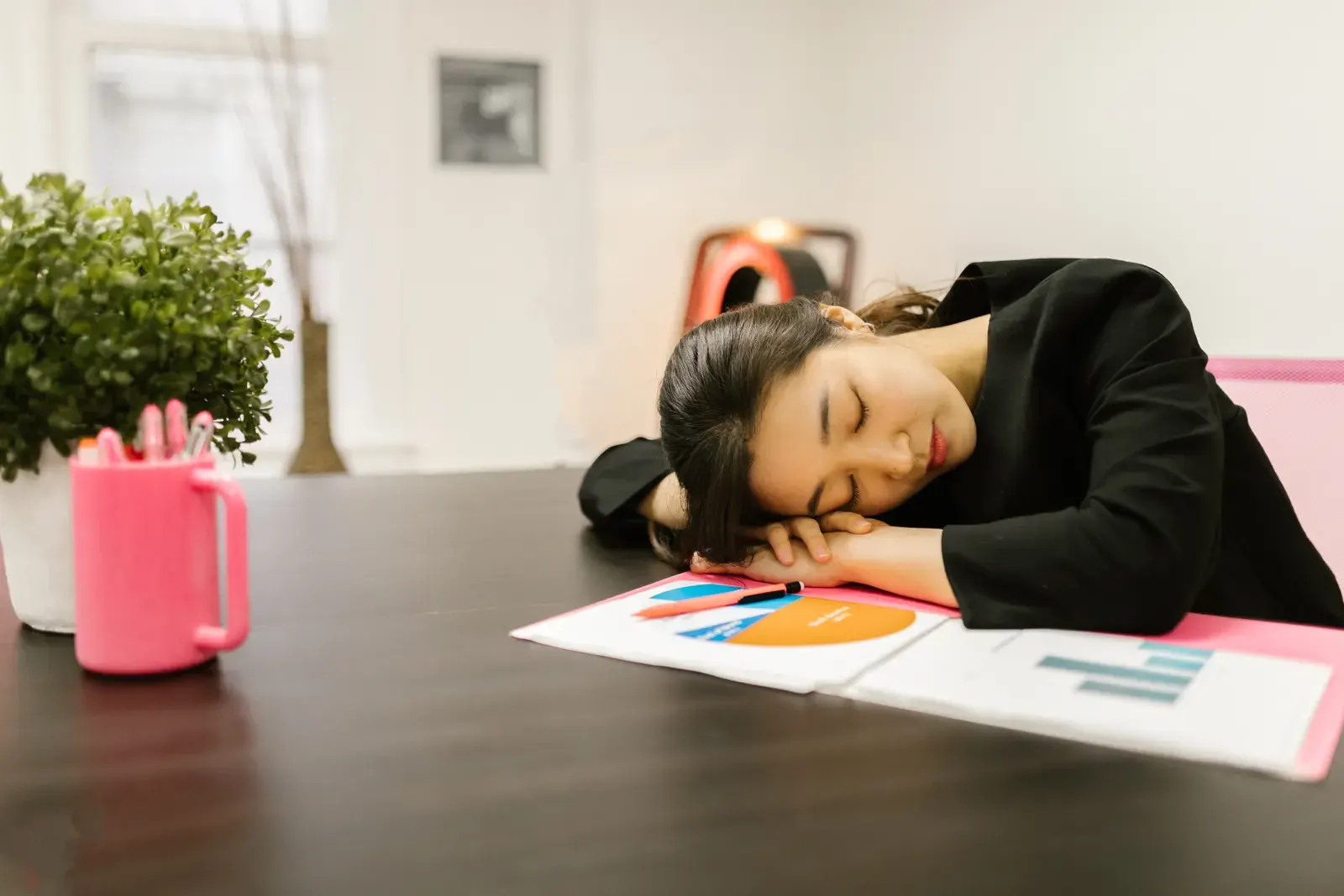 An exhausted woman in a black outfit rests her head on a desk in an office setting, surrounded by papers and a potted plant.