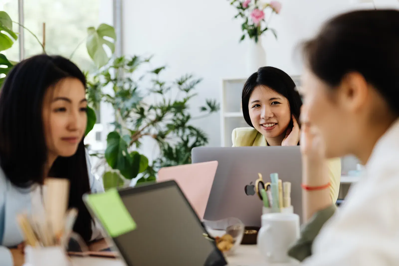 Three women in a modern office setting discussing work with laptops.