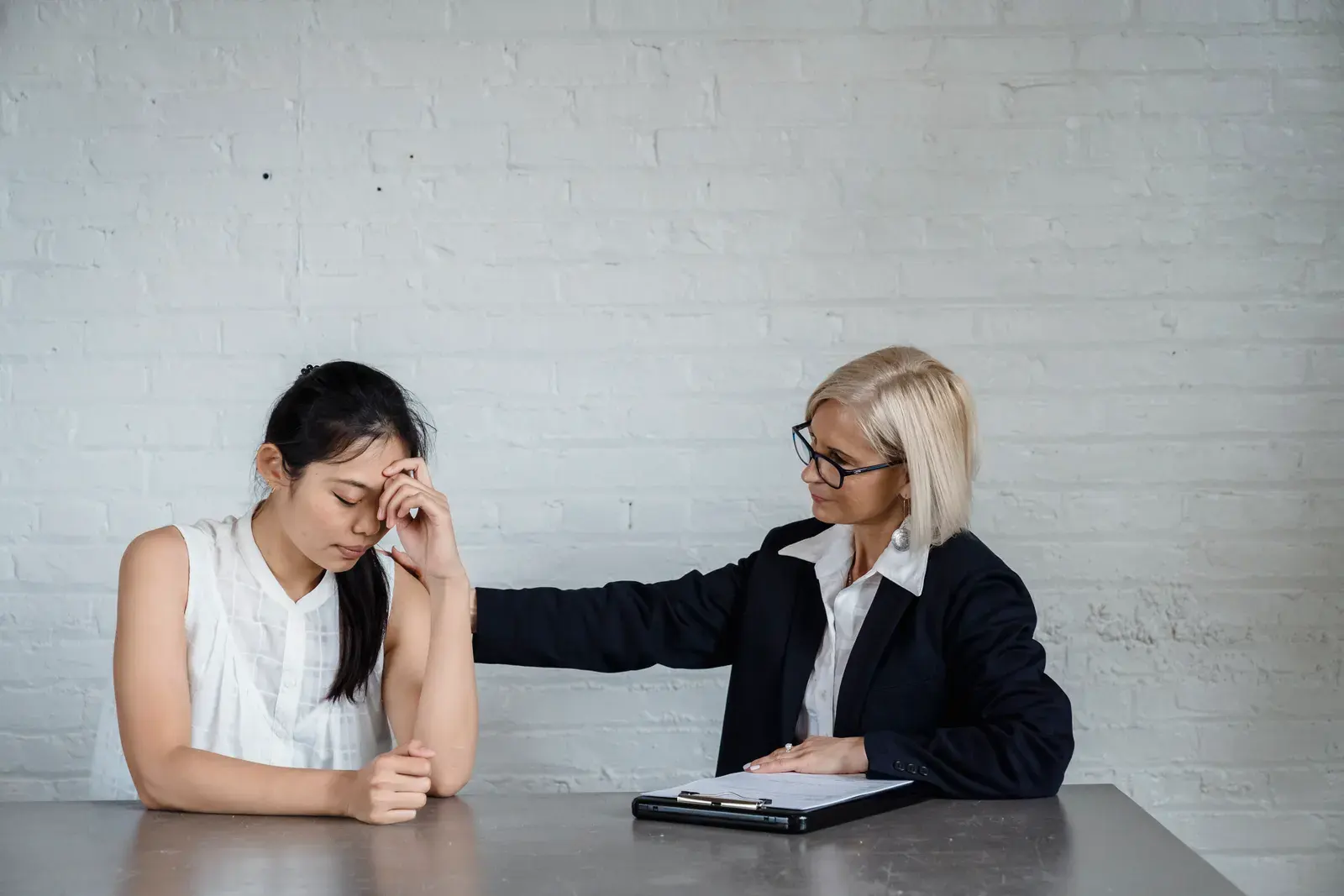 A supportive counseling session where one woman comforts another at a table against a brick wall.