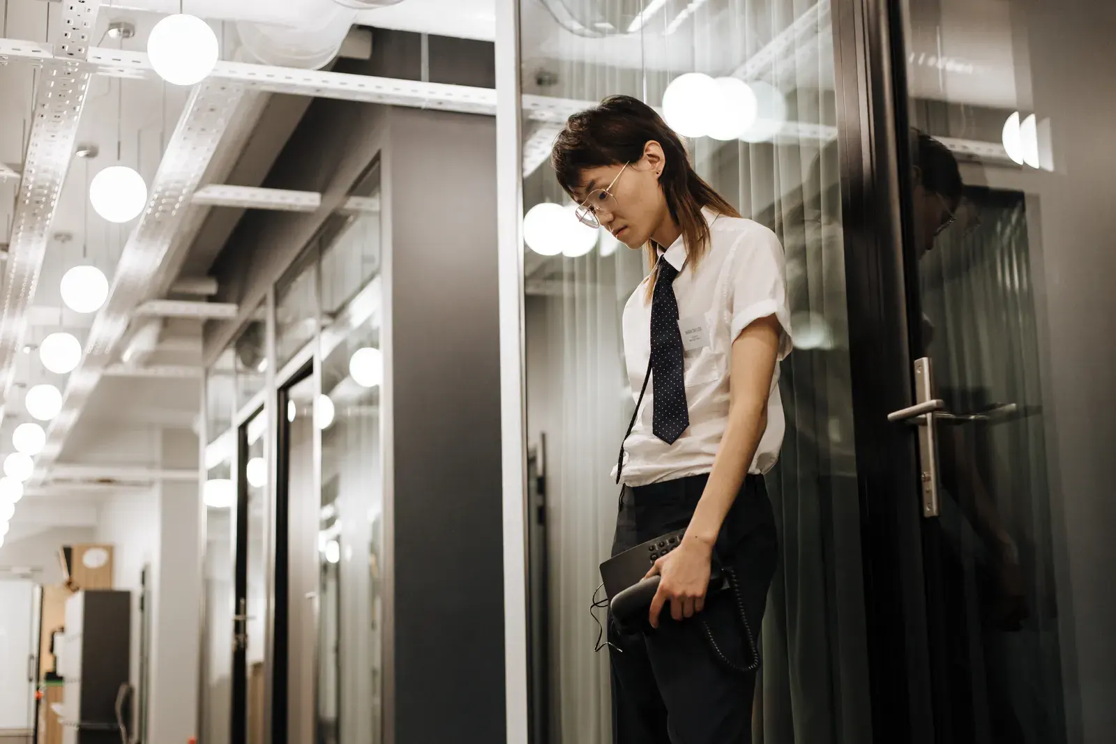 Young Asian adult leaning against a glass door in an office, appearing stressed, holding a tablet.