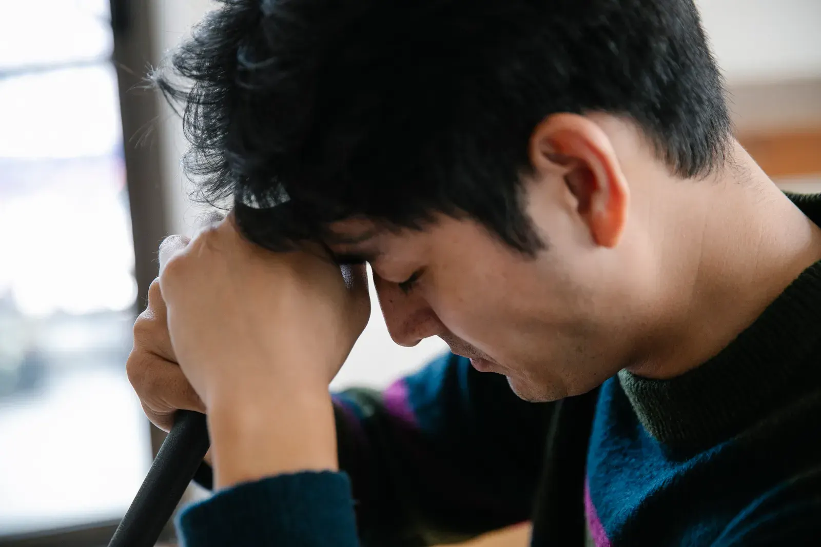 Close-up of a man looking stressed as he rests his head on his arms indoors.