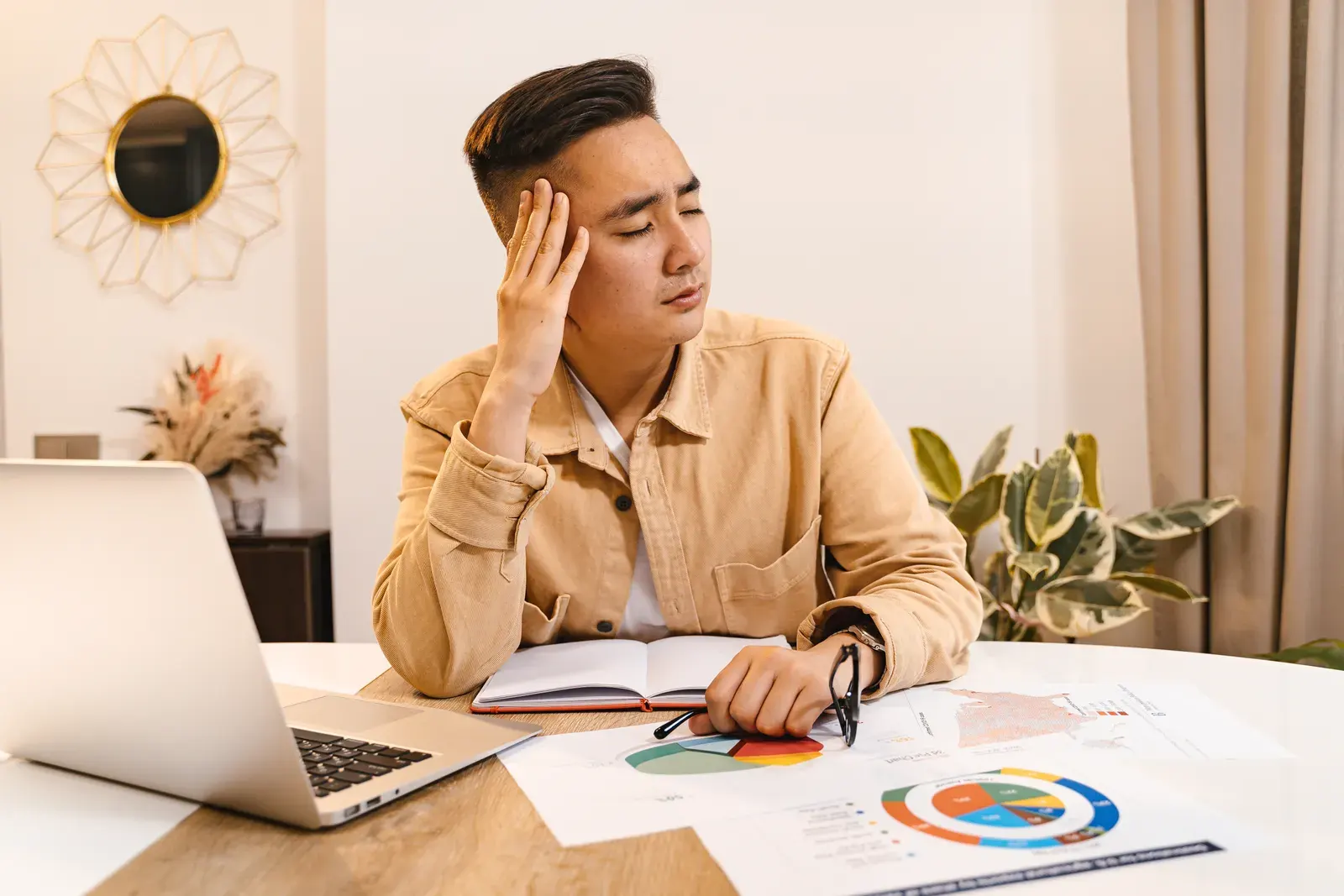 Businessman holding head in frustration, surrounded by documents and laptop at home office.
