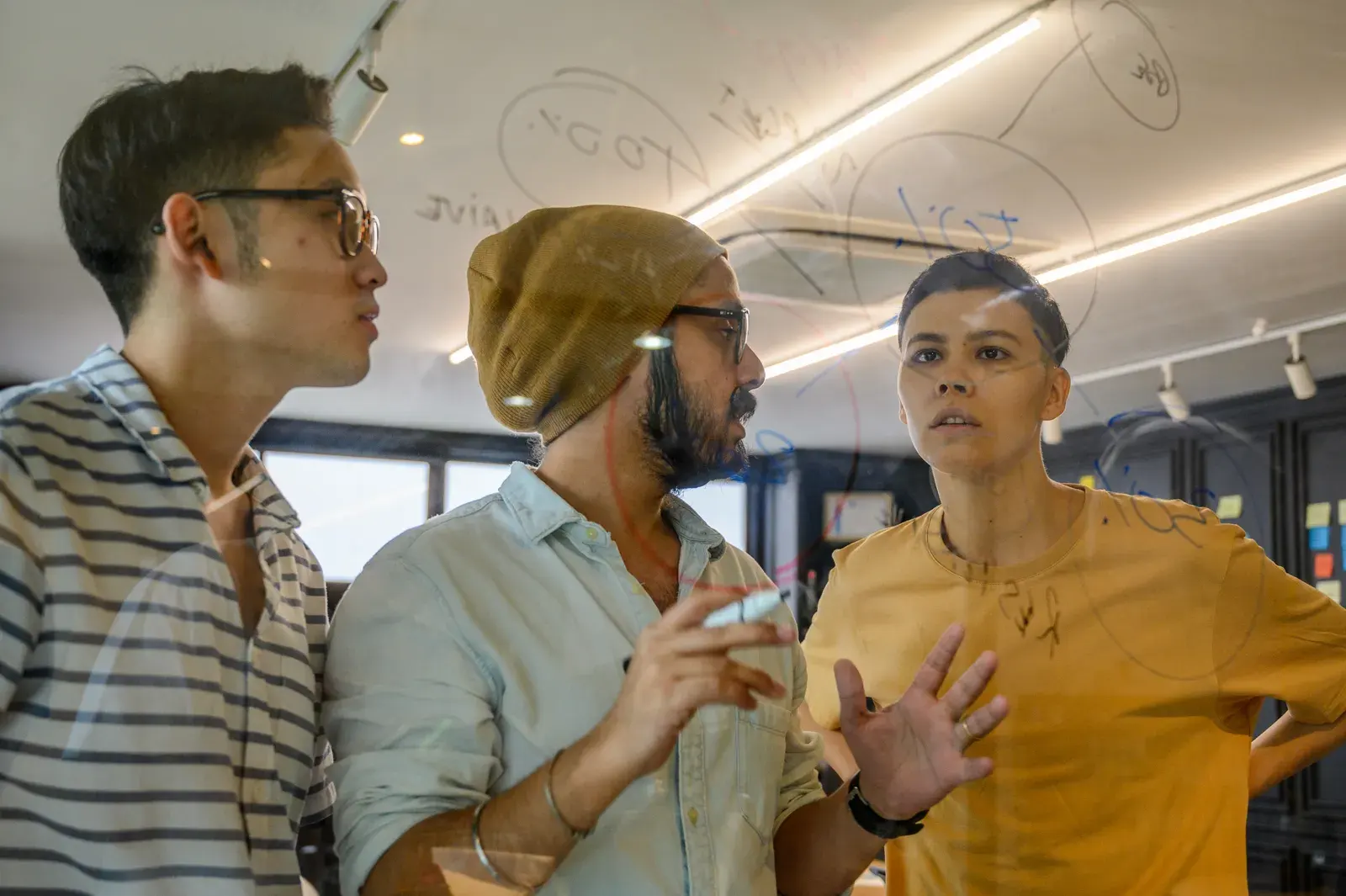 A diverse group of young professionals brainstorming and writing ideas on a glass board in a modern office setting.