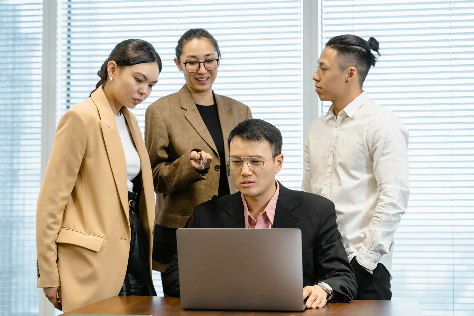 Group of colleagues in formal wear actively discussing work around a laptop in office setting.