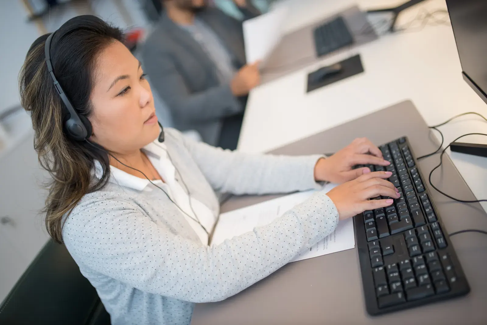 Asian woman working at a computer with headphones in a modern office setting.