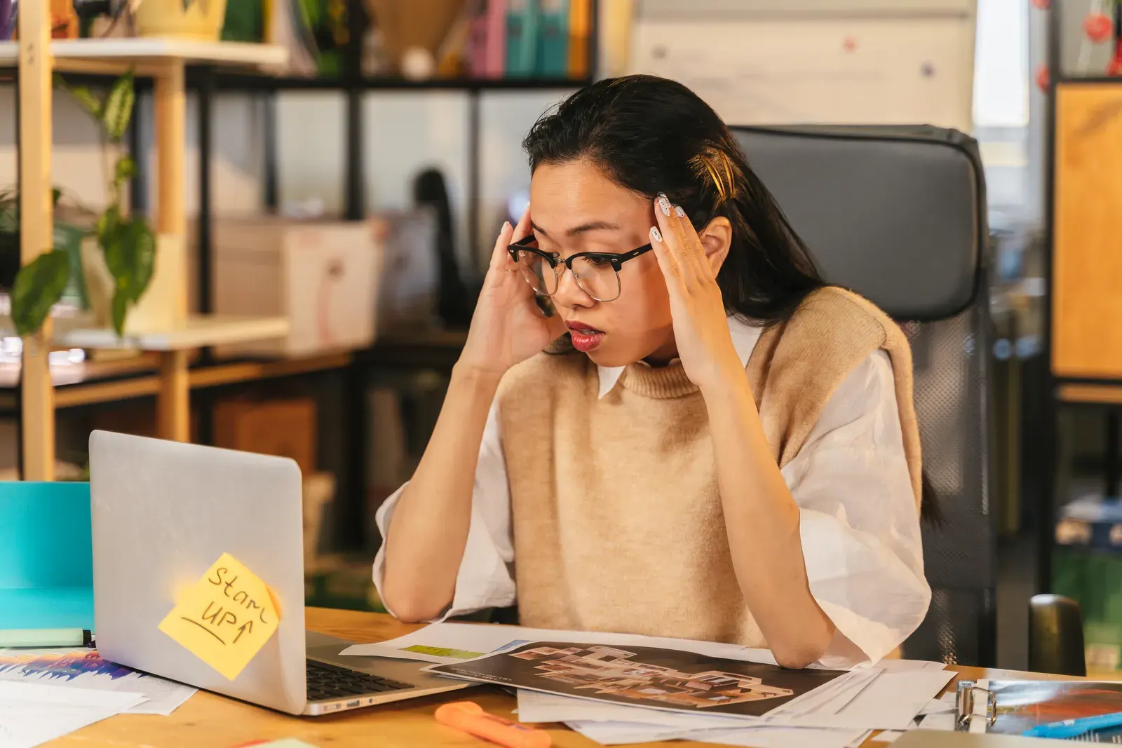 A woman with glasses shows stress while working at a laptop in an office environment.