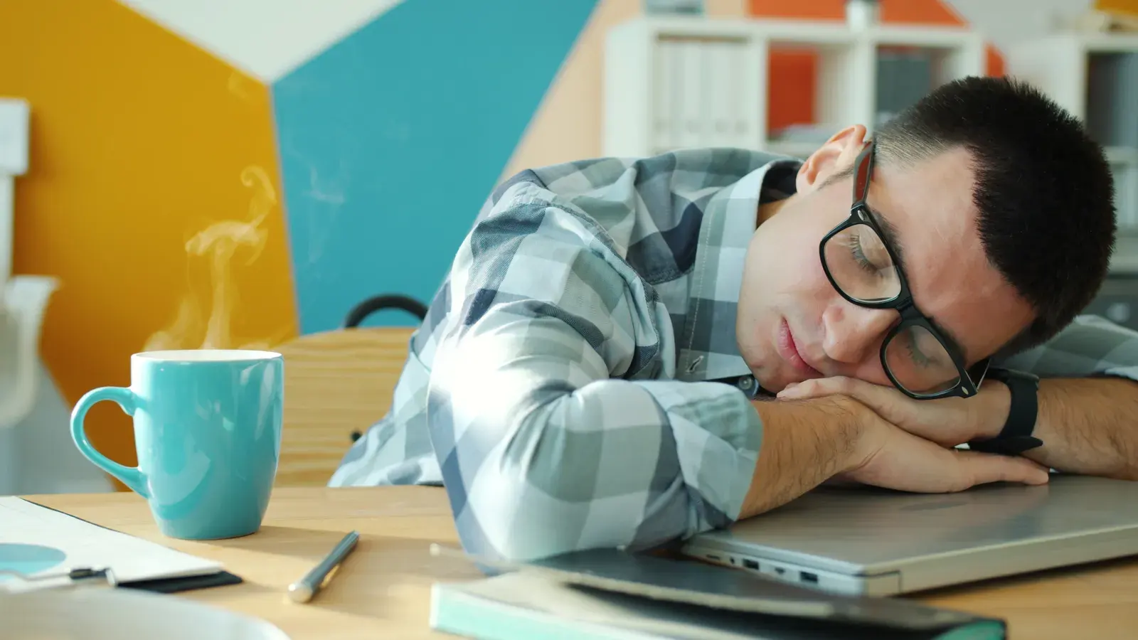 Tired man sleeping on laptop with coffee, showcasing workplace fatigue.