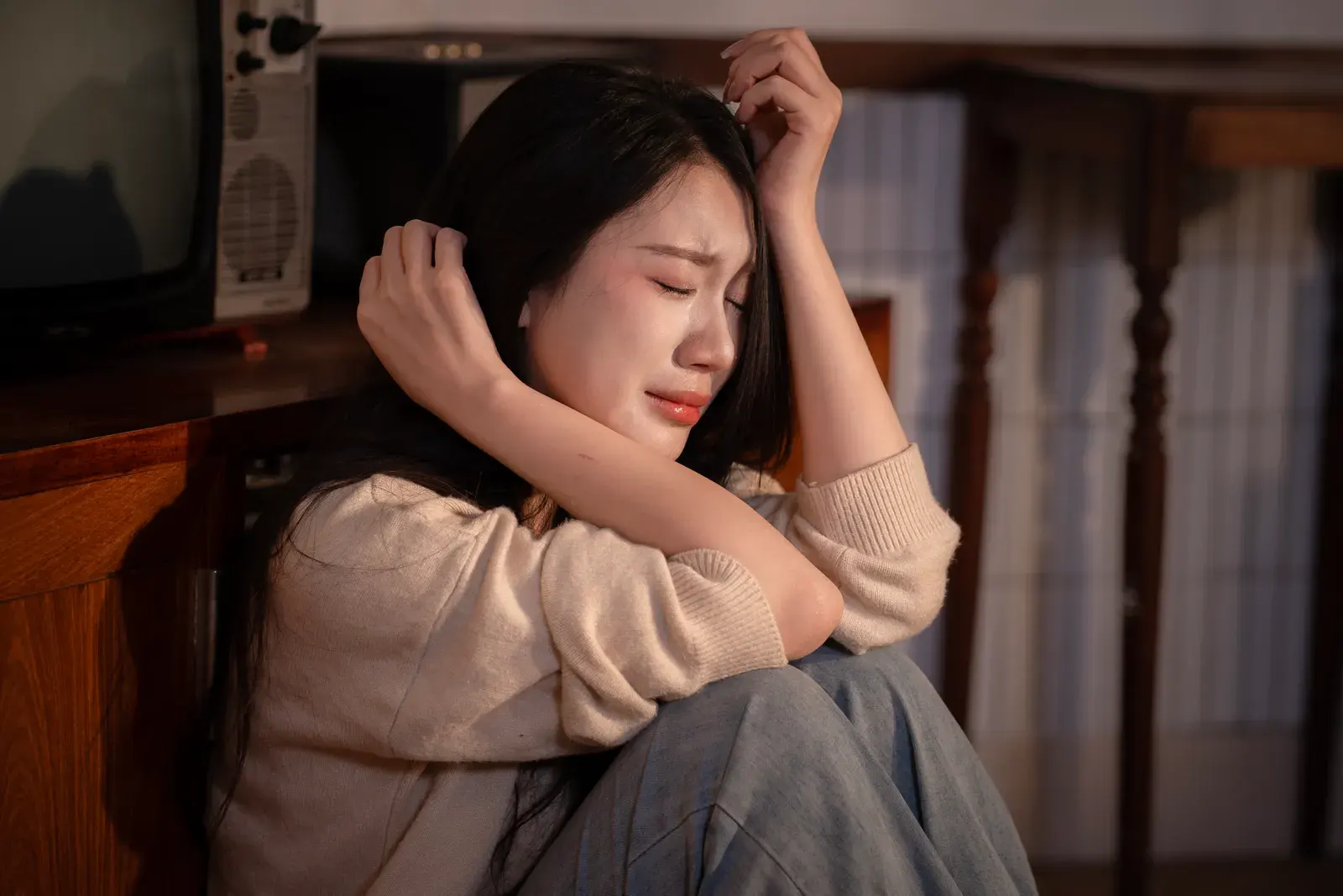 A young woman sitting indoors, expressing deep emotional distress.