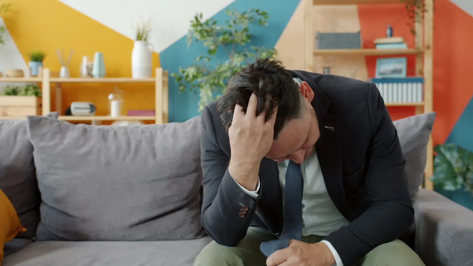 A worried businessman sits indoors on a colorful couch, holding his head in stress.